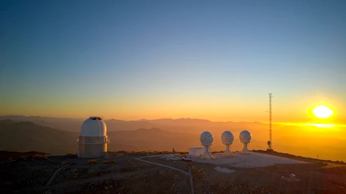 Eine große Teleskopkuppel links und drei kleinere Kuppeln auf einer Art Ständer nebeneinander rechts auf einem Berggipfel im Licht der tiefstehenden Sonne