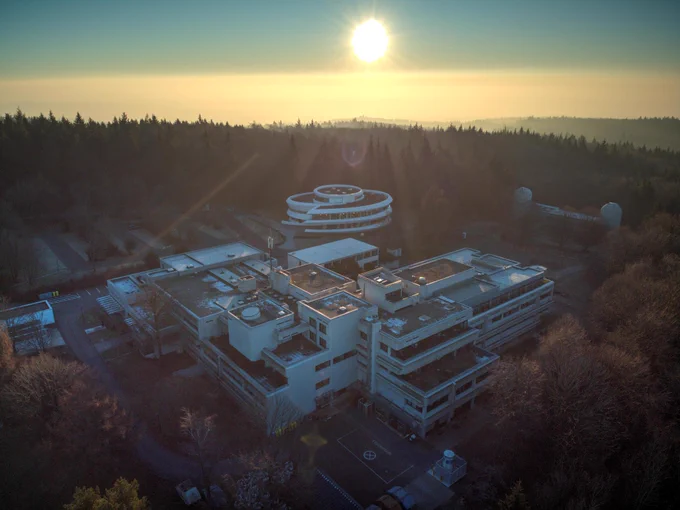 Drohnenaufnahme des MPIA-Campus mit tiefstehender Sonne über einer Nebelschicht, Lichtstrahlen gehen durch die Bäume