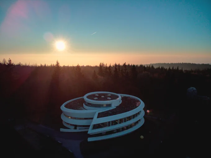 Drohnenaufnahme des Hauses der Astronomie mit tiefstehender Sonne über einer Nebelschicht, Lichtstrahlen gehen durch die Bäume