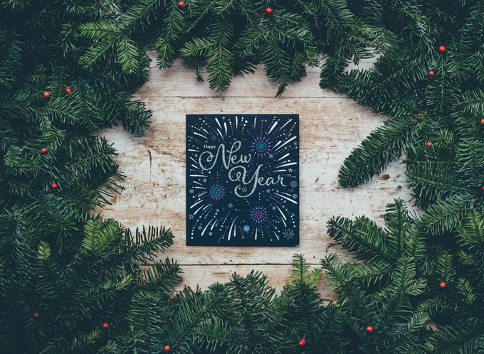 A black, squared, board with the text "Happy New Year" written in silver sparkling text. The text is surrounded by painted fireworks in pink, purple, green, blue and silver. The background is made up by light wooden board with a frame of green pine branches with red berries. Photo by Annie Spratt on Unsplash