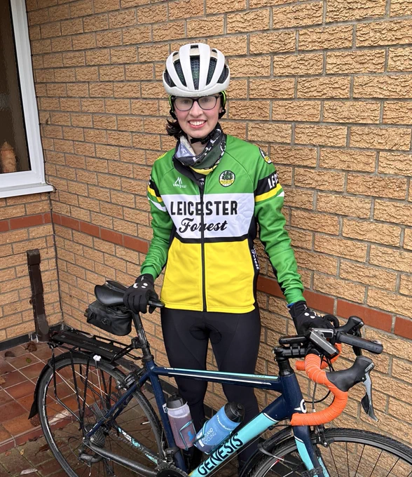 A woman in a white helmet, green and yellow jacket and black trousers holding a blue road bike in front of her. There is a brick wall in the background. There are drops of water on her helmet and her glasses are steamed up.