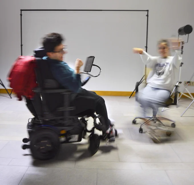 Un chico da vueltas rápidamente con su silla de ruedas. Enfrente, una chica da vueltas, con las manos extendidas hacia arriba, en una silla de oficina con ruedas. Él viste de oscuro, y ella de gris claro. La fotografía realizada en un local diáfano, da la sensación de estar viendo un baile.