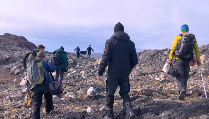 A group of people dressed in outdoor gear and backpacks hike across a rocky, rugged landscape in Greenland under a clear sky. Two individuals in the distance are carrying a black container. The terrain consists of scattered rocks and patches of sparse vegetation.