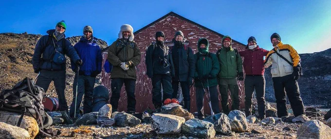 A group of nine people in warm, layered clothing and outdoor gear pose in front of a red wooden building in a rocky, remote area of Greenland. The group is standing on uneven terrain with scattered boulders under a clear blue sky.