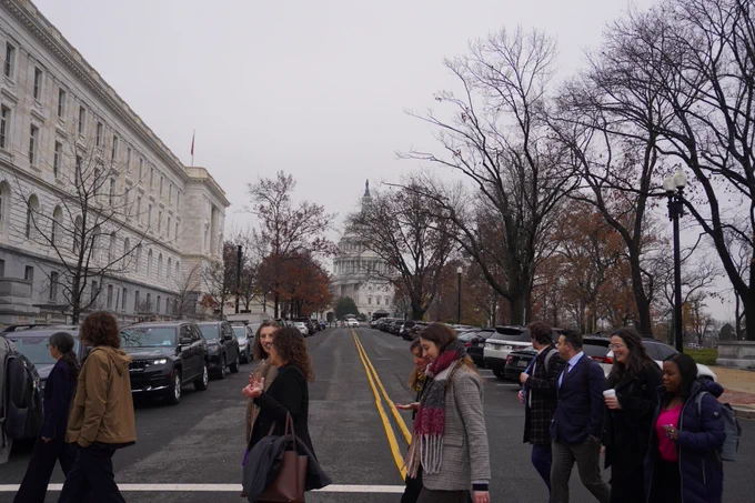 A group of people walk across the street in the U.S. Capitol.  The U.S. Capitol building can be seen in the background against a grey sky. 
