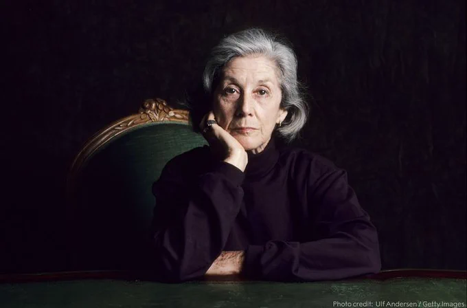 Nadine Gordmier, a grey haired woman, looking directly into the camera. She has her chin in her right hand. She is wearing a black shirt and sitting on a green chair.