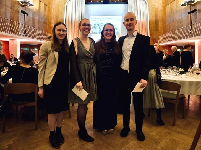 Four early-career researchers pose together at a formal event in an elegant hall with wooden panels and a balcony. 