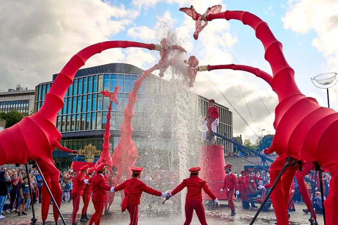 Performers dressed in red uniforms interact with large red inflatable giraffe puppets at a fountain in City Park. There is a crowd of onlookers in the background.