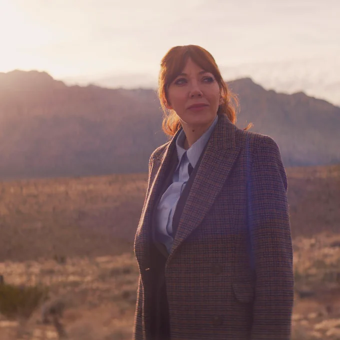 Philomena Cunk stands outdoors against a backdrop of mountains, wearing a suit and looking contemplative as she gazes at something off camera