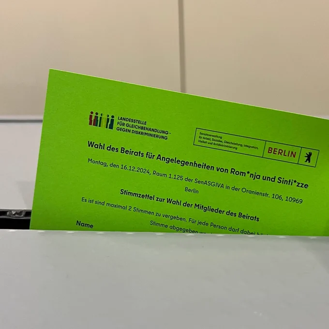 Ein grüner Wahlzettel wird in die Wahlurne eingeworfen. Darauf stehen die Logos der Landesstelle für Geleichbehandlung - gegen Diskriminierung und der Senatsverwaltung für Arbeit, Soziales, Gleichstellung, Integration, Vielfalt und Antidiskriminierung.
Die Überschrift:
"Wahl des Beirats für Angelegenheiten von Rom*nja und Sinti*zze 
Montag, den 16.12.2024, Raum 1.125 der SenASGIVA in der Oranienstr. 106, 10969
Stimmzettel zur Wahl der Mitglieder des Beirats"