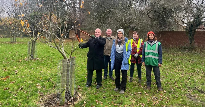 A group of people standing around a young fruit tree in an orchard in winter, smiling at the camera. One person is holding a pruning tool.