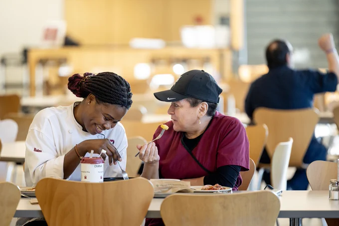 Bates Dining Services staff members Diana Mba Oyana (left, wearing white) and Sonia Roy (right, wearing garnet and a black baseball cap) finished their early dinner before the arrival of the supper crowd, they enjoyed a quiet moment. Or maybe a private joke.