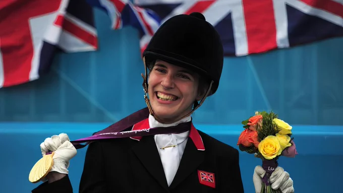 Sophie dressed in a competition jacket and riding hat holding a gold medal from London 2012 and a bouquet of flowers. Behind her are union jack flags.