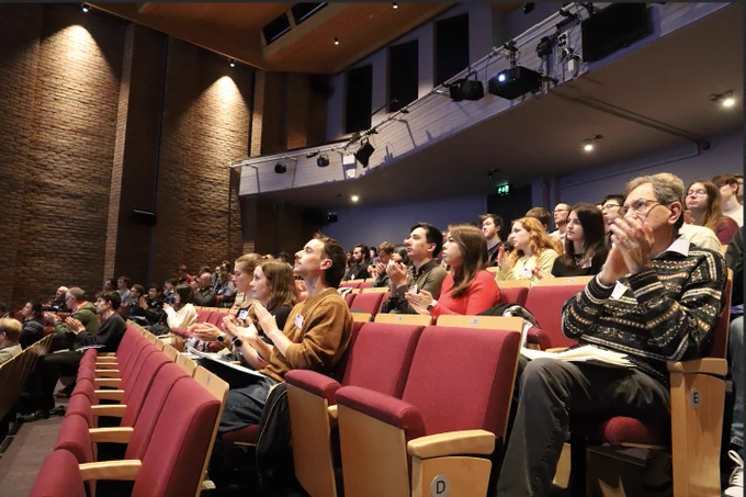 audience in the lecture theatre clapping. The lecture theatre is dark and ornate with low, warm lighting and red and wooden seats.