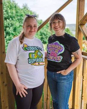 Two people standing side by side, one wearing a white t-shirt that reads "Sobriety is not superiori" the other is wearing a black t-shirt that reads "Patriarchy Got Me Drove."