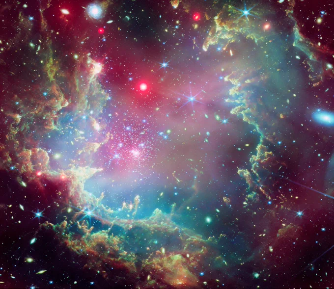 This is a composite image of star cluster NGC 602, in vibrant and festive colors. The shape of the star cluster strongly resembles a holiday wreath. The cluster includes a giant dust cloud ring, shown in greens, yellows, blues, and oranges. The green hues and feathery edges of the ring cloud create the appearance of a wreath made of evergreen boughs. Hints of red representing X-rays provide shading, highlighting layers within the wreath-like ring cloud.

The image is aglow with specks and dots of colorful, festive light, in blues, golds, whites, oranges, and reds. These lights represent stars within the cluster. Some of the lights gleam with diffraction spikes, while others emit a warm, diffuse glow. Upon closer inspection, many of the glowing specks have spiraling arms, indicating that they are, in fact, distant galaxies. Credit: X-ray: NASA/CXC; Infrared: ESA/Webb, NASA & CSA, P. Zeilder, E.Sabbi, A. Nota, M. Zamani; Image Processing: NASA/CXC/SAO/L. Frattare and K. Arcand