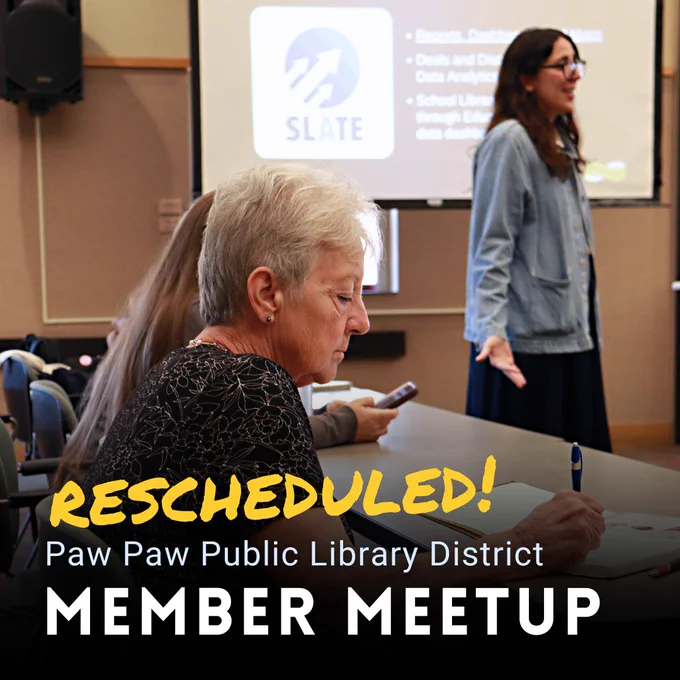 A person sits at a table and writes in a notebook. A person in the background speaks to an audience. Rescheduled: Paw Paw Public Library District Member Meetup. 