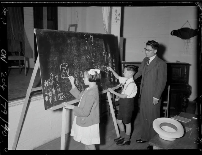 A black and white photograph depicting a man in a pinstripe suit looking on as two children write in Chinese on a blackboard. 'Adults and children at Chinese lessons (January 1945)', Mitchell Library, State Library of New South Wales. Accessed through the OMAA Archive.