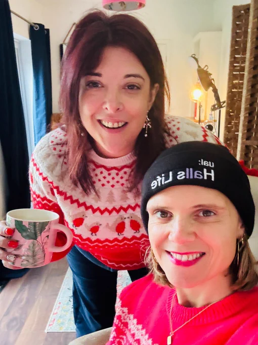 Two women are in a selfie photo smiling at the camera. One has brown hair and a Christmas jumper on. she is holding a pink mug. The other has blonde hair and is wearing a black beanie hat and a red Xmas jumper