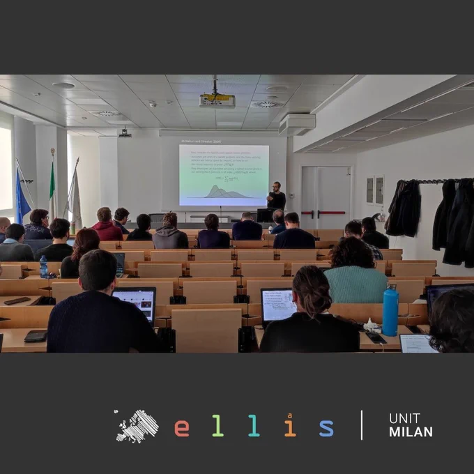Presentation at ELLIS Unit Milan, featuring an individual speaking in front of an audience in a classroom with laptops, seated at wooden desks.