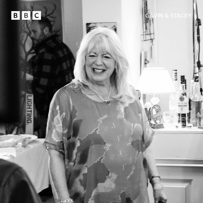 Alison Steadman (Pam) smiling brightly while standing in a cozy indoor set. Behind her, a bar area with bottles can be seen.