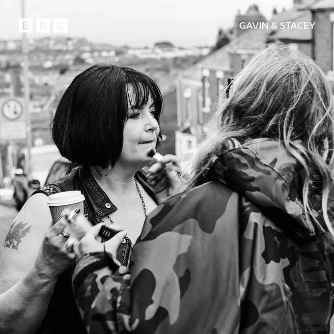 Ruth Jones (Nessa) getting a touch-up on her makeup from a crew member. She holds a coffee cup in one hand, her iconic leather vest visible as she stands on set.