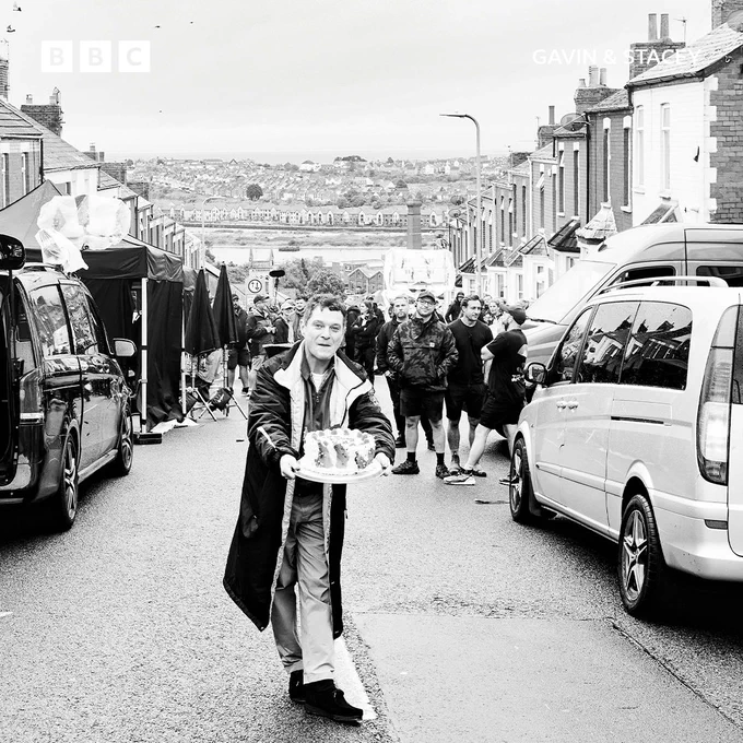 Mathew Horne (Gavin) holding a decorative cake, walking down a lively residential street filled with crew and equipment.