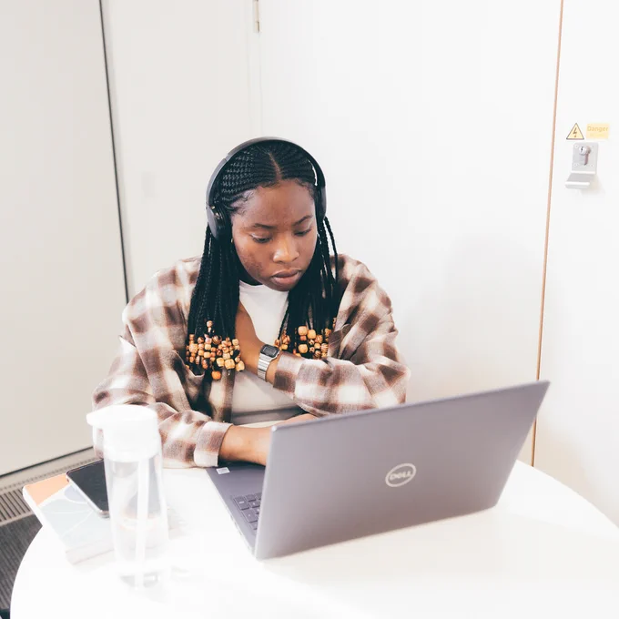 A student wearing headphones sits at a round library table. She has a water bottle next top her and is typing on a laptop.