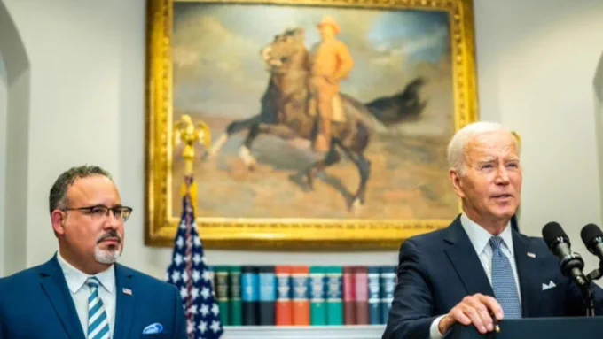 U.S. President Joe Biden speaking at a podium with another individual standing beside him, in a room with a painting of a horseback rider in the background and American flags.