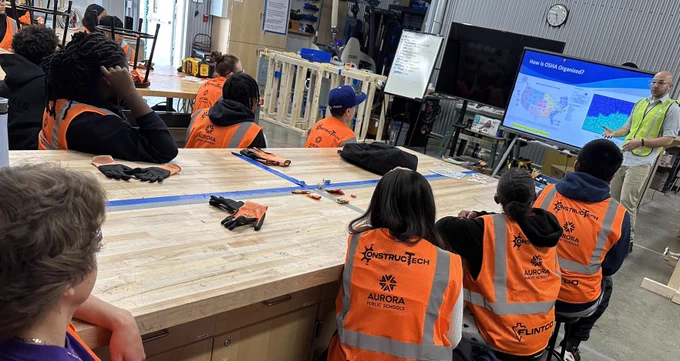 A group of high school students in orange safety vests with "ConstructTech" logos are attentively listening to a presenter in a workshop area. The presenter, standing next to a digital screen displaying a map, is explaining points of interest. Various tools are scattered on the worktable in front of the attendees.