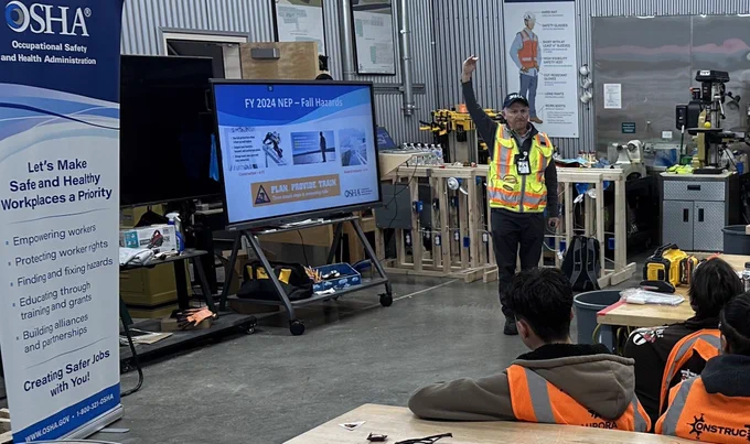 A person wearing safety gear including a high-visibility vest is presenting at an Occupational Safety and Health Administration (OSHA) workshop in a workshop setting, surrounded by attentive attendees in similar safety attire. There are industrial tools and workbenches in the background.