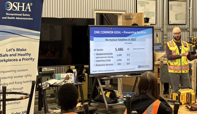 Person presenting workplace safety statistics for 2022 next to a screen displaying data, in a workshop setting with an OSHA (Occupational Safety and Health Administration) banner visible.