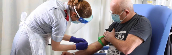 A nurse stands by a patient sat in a chair, preparing to take blood from the patient's arm