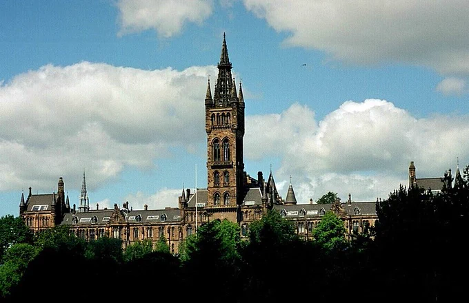 View of the yellow sandstone Glasgow University tower and its wings extending either side below. The architectural style is Gothic Revival. The photo is taken from across the River Kelvin close to Kelvingrove Art Gallery & Museum. 