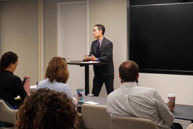 A professionally dressed man presents to a group of attendees in a conference room setting. The presenter is standing behind a podium. Several audience members are visible, seated and facing the presenter.
