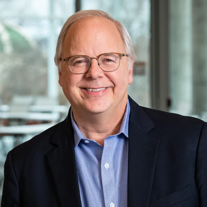 Michael Trick wearing glasses, a blue blazer, and a blue shirt, standing indoors with large windows and blurred greenery in the background.