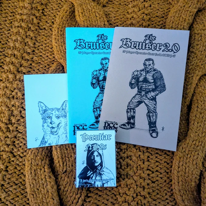 a white postcard of a canine. 

a blue and a kraft-brown copy of The Bruiser 2.0 mini zines. featuring a brute of a man with metal gauntlets. 

the box of Peculiar NPCs, a deck of fantasy character portraits.

on an orange knit blanket.