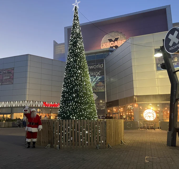 A person dressed as Santa Claus stands next to a large, illuminated Christmas tree in front of the Xscape building during twilight.