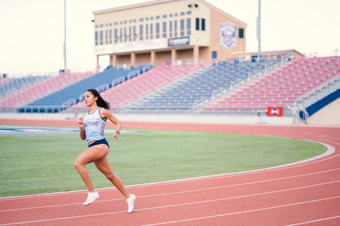 Colorado State University Pueblo alum Yasmine Hernandez runs on a red track on the university's campus. She is wearing a gray track-and-field jersey that reads "Timberwolves," navy bottoms and white track spikes. Red and blue stands, a tan-colored press box and a green space in the center of the track compose the background of the photo. 