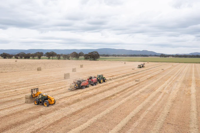 Aerial view of an agricultural field with three tractors working, hay bales scattered across the landscape, and mountains in the background under a cloudy sky. The AgriFutures #ExportFodder Program is seeking a qualified provider to conduct a detailed evaluation of the current annual #ryegrass toxicity assay and the development of alternative assays and guidance on potential changes to the current sampling system.