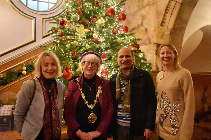 Four people stand in front of a large Christmas tree covered in lights and decorations in Wigmore Hall, facing the camera and smiling. Second from the left is the Deputy Lord Mayor of Westminster wearing her civic regalia, a large golden necklace.