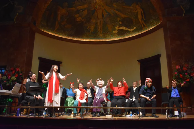 Around 15 choir members are sat on chairs on the stage at Wigmore Hall with their hands in the air. A female choir leader stands to the left of the stage facing the camera.