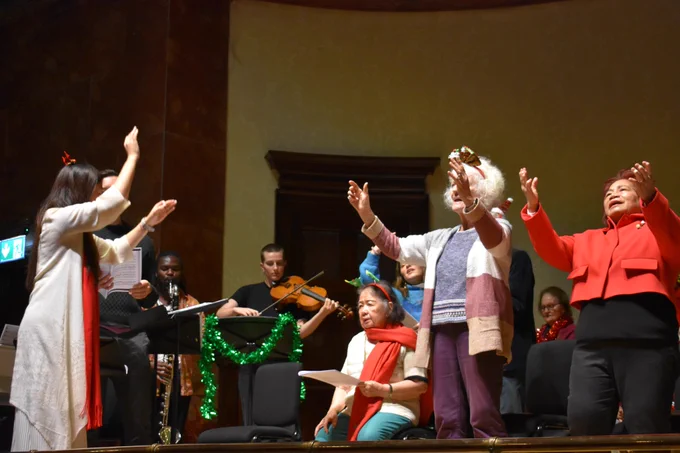 3 choir members are stood up singing with their hands in the air and another 3 members are sat on chairs singing on the stage at Wigmore Hall. A female choir leader stands to the left of the stage with her hands in the air conducting.