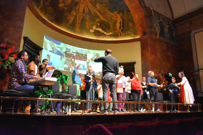 Around 15 choir members are sat on chairs on the stage at Wigmore Hall with their hands in the air. To the left of the picture we can see a pianist, a bass clarinettist and someone playing a violin. Behind the choir on the back wall of the stage is a video projection of a Zoom call featuring 6 different screens of online choir members singing along.