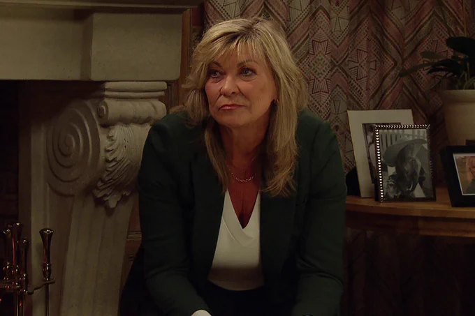 A triumphant looking Kim Tate in Emmerdale sitting down and leaning forward, wearing a green blazer and white top.