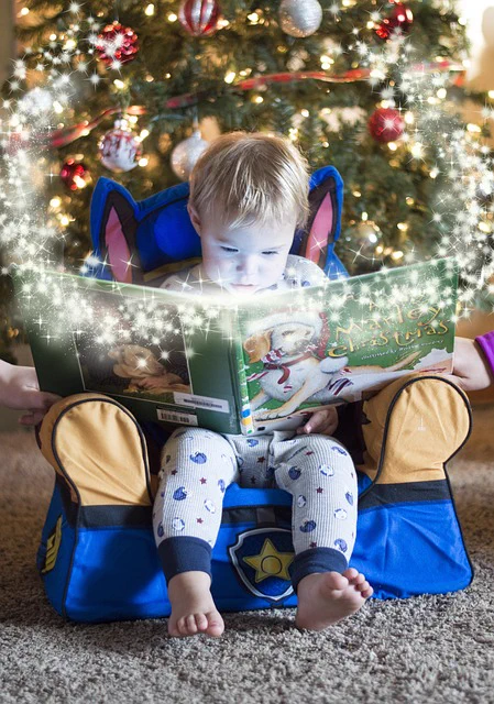 A child sits on a small chair under a Christmas tree, reading a large picture book about Christmas.