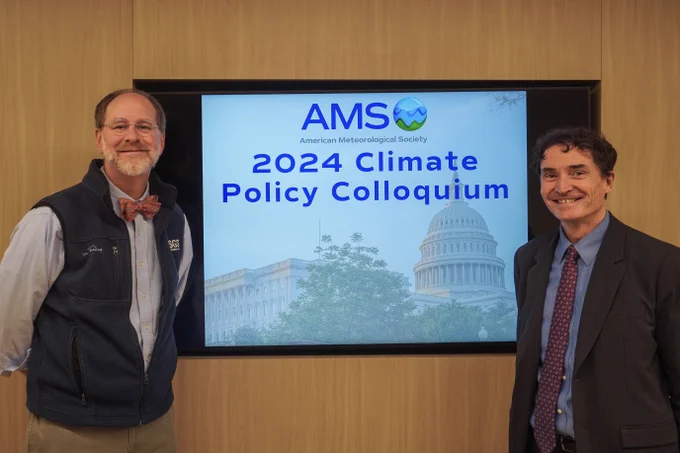 Two men stand beside a presentation screen displaying "AMS 2024 Climate Policy Colloquium" with the American Meteorological Society logo and an image of the U.S. Capitol.