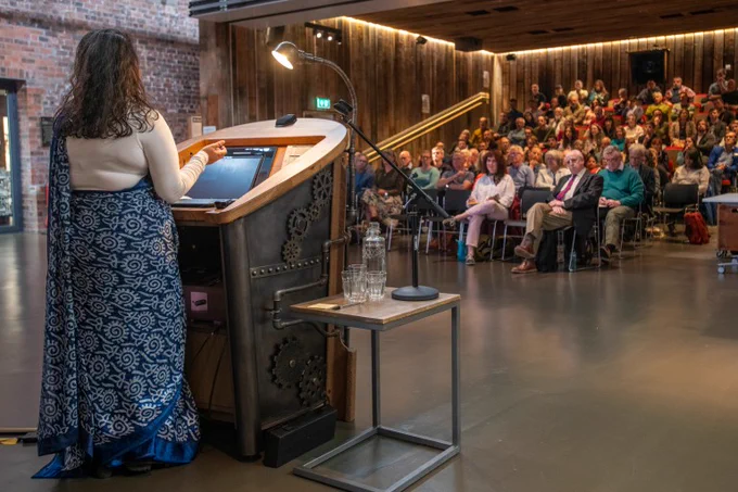 A person wearing a long, patterned skirt, standing at a lectern speaking to a large group of people seated inside the Engine Shed