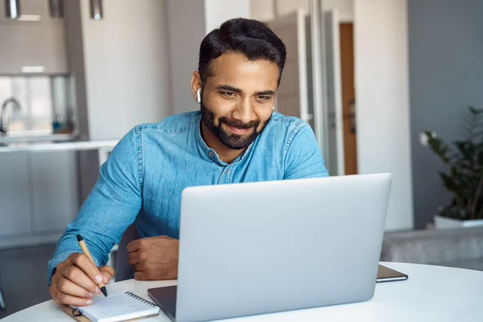 Stock image of a man sitting at a computer, smiling as he is writing with a pen on a notepad.
