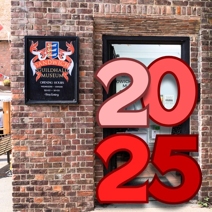 A photograph of Sandwich Guildhall Museum's entrance, with a black sign that has a red, yellow and blue coat of arms and text reading "Sandwich Guildhall Museum, opening hours, Wednesday to Sunday, 10:00 to 16:00, free entry." In the door way of the museum there are edited numbers, which are read, reading 2025, with 20 on the top line and 25 on the lower line.